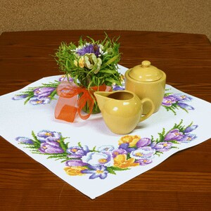 Pu&ograve; includere: Una tovaglia bianca con un ricamo floreale che presenta crochi viola e gialli. Due contenitori per panna e zucchero in ceramica gialla sono sulla tovaglia, insieme a un mazzo di fiori.