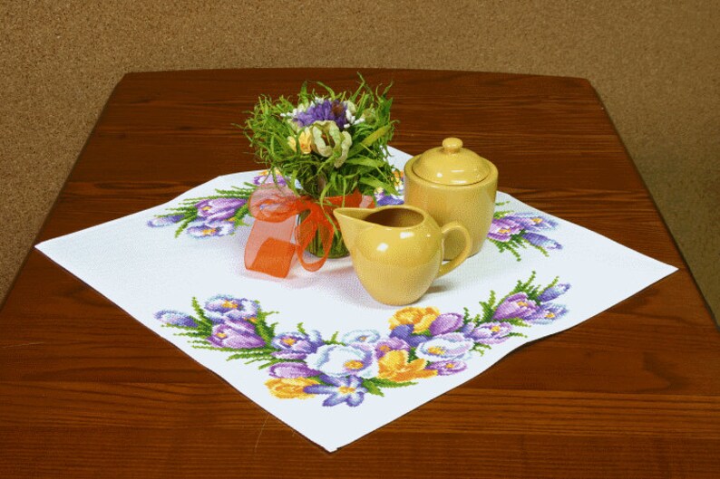 Pu&ograve; includere: Una tovaglia bianca con un ricamo floreale che presenta crochi viola e gialli. Due contenitori per panna e zucchero in ceramica gialla sono sulla tovaglia, insieme a un mazzo di fiori.