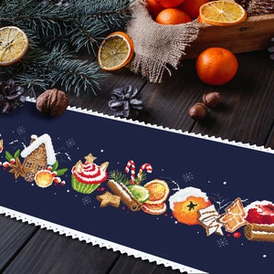Puede incluir: Un camino de mesa azul con un borde de encaje blanco presenta un diseño navideño festivo con casas de jengibre, cupcakes, palitos de canela, naranjas y manzanas rojas.