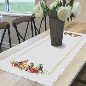 Puede incluir: Camino de mesa rectangular blanco con un diseño floral y de pájaros. El diseño bordado presenta tres pájaros posados en una rama con flores rojas y amarillas. El camino de mesa tiene un borde de encaje festoneado y un borde dorado.