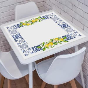 Puede incluir: Mantel cuadrado blanco con un diseño de borde floral azul y blanco. El borde presenta un patrón repetitivo y acentos de limón y flores. El mantel está sobre una mesa blanca con sillas blancas.