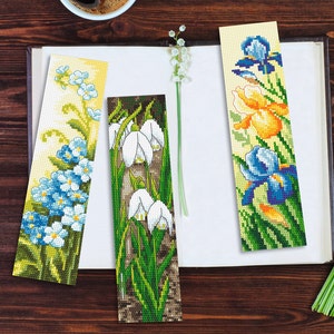 May include: Three cross-stitch bookmarks featuring floral designs. The first bookmark has blue forget-me-not flowers, the second has white snowdrops, and the third has blue and yellow irises.