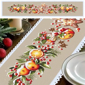 Puede incluir: Un camino de mesa beige con un borde de encaje blanco presenta un diseño navideño festivo. El diseño incluye naranjas, bayas de acebo, ramas de pino y galletas de jengibre.