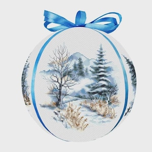 May include: A circular cross-stitch decoration depicting a winter scene with snow-covered trees and mountains in blue and grey hues. A blue ribbon is attached at the top. The ornament is approximately 10 cm in diameter.