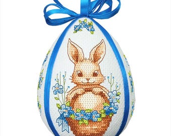 Può includere: Un uovo di Pasqua bianco con un nastro blu e un coniglietto marrone che tiene un cestino di fiori blu.