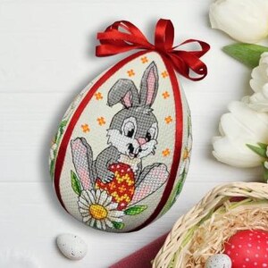 Peut inclure: Décoration en forme d'œuf de Pâques avec un nœud en ruban rouge. L'œuf présente un motif au point de croix d'un lapin gris tenant un œuf décoré, entouré de fleurs. L'œuf est sur une surface blanche avec des fleurs blanches et un panier.