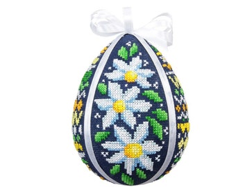 Può includere: Un ornamento decorativo a forma di uovo con sfondo blu scuro, con un disegno a punto croce di margherite bianche con centri gialli e foglie verdi. Un nastro bianco è attaccato per appenderlo. Una decorazione pasquale unica.