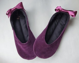 May include: A pair of plum-colored velvet slippers with a satin bow on the heel. The slippers have a dark interior and a rounded toe. The brand name is visible in gold lettering inside the slipper.