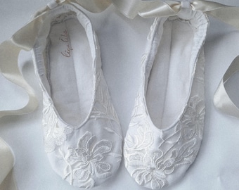 Puede incluir: Un par de zapatillas de ballet blancas con bordados florales. Las zapatillas tienen cintas de raso para atar y el interior está forrado con tela blanca. Las zapatillas son un diseño delicado y femenino.