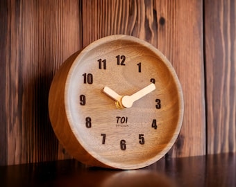 Puede incluir: Un reloj redondo de madera con una esfera clara y números marrón oscuro. Las manecillas del reloj son claras y la esfera tiene el texto "TOI DESIGN". El reloj está sobre un fondo de madera.
