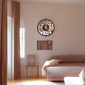 May include: A light brown wooden clock with Roman numerals and a white face hangs on a light pink wall. The clock is in a living room with a light brown couch and a small side table.