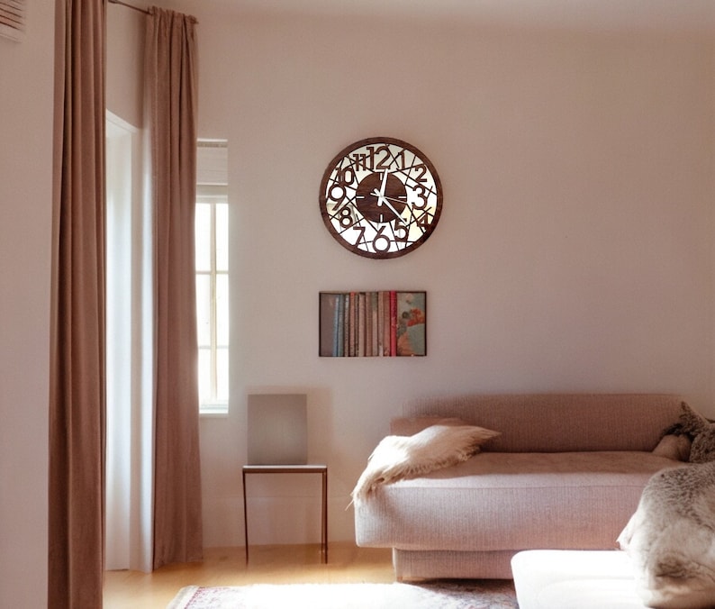 May include: A light brown wooden clock with Roman numerals and a white face hangs on a light pink wall. The clock is in a living room with a light brown couch and a small side table.