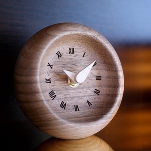 Op de afbeelding: Een houten klok met Romeinse cijfers op de wijzerplaat. De klok is gemaakt van donkerbruin hout en heeft een witte wijzer.