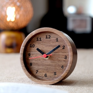 May include: A wooden clock with a dark brown finish and black hands. The clock face has white numbers and a red second hand. The clock is sitting on a brown surface.