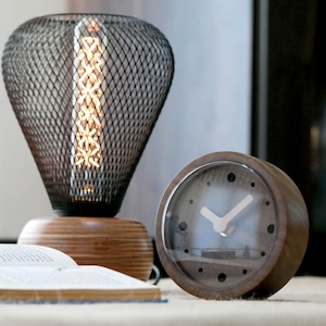 May include: A brown wooden clock with a white face and black hands. The clock is sitting on a table next to a black metal lamp with a light bulb inside.