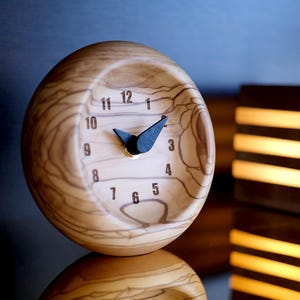 May include: A wooden clock with black hands and Roman numeral markings. The clock is round and has a wood grain pattern. It is sitting on a dark surface.