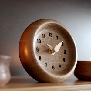May include: A wooden desk clock with a light-colored face and black numerals. The clock has a curved design and light-colored hands. Two lit candles in brown holders flank the clock. The clock is on a wooden surface.