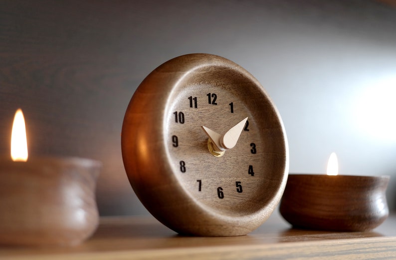 May include: A wooden desk clock with a light-colored face and black numerals. The clock has a curved design and light-colored hands. Two lit candles in brown holders flank the clock. The clock is on a wooden surface.