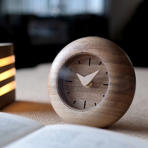 May include: A wooden clock with a round face and a light brown finish. The clock has a white hour hand and a black minute hand. The clock is sitting on a table with a book and a wooden lamp.