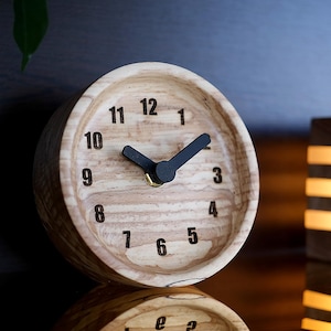 May include: A wooden clock with black hands and a light wood grain finish. The clock face has black numbers 1 through 12.