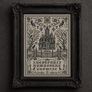 May include: A black and white cross-stitch sampler in a dark ornate frame. The design features a central gothic mansion, bare trees, crows, and roses. Below the house is the full alphabet in a gothic font, surrounded by decorative floral borders.