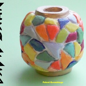 May include: A small, round, ceramic vase with a mosaic design. The vase is decorated with colorful, geometric shapes in shades of red, orange, yellow, green, blue, and purple. The vase has a gold rim and is sitting on a light green surface. The text "Potent Assemblage" is written below the vase.