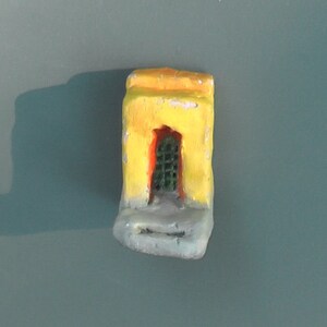 May include: A small, hand-painted, yellow and gray miniature building with a green arched doorway. The building has a weathered appearance, with visible brushstrokes and paint chipping. The doorway features a green, grid-like pattern.