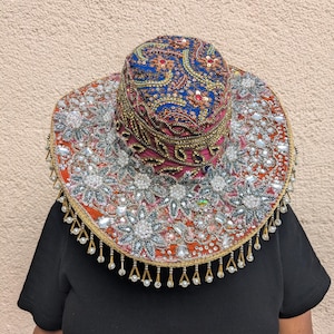 Könnte beinhalten: Ein breitkrempiger Hut mit einer blauen Krone, die mit aufwendigen goldenen und roten Stickereien sowie Pailletten verziert ist. Die Krempe ist orange und mit silbernen Blumenperlen, klaren Kristallen und weißen Perlenfransen besetzt.
