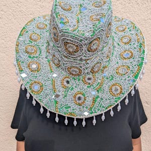 May include: A green hat featuring intricate silver and gold beadwork, circular patterns, and embedded mirror pieces. The brim is adorned with a fringe of small white teardrop-shaped beads hanging from the edge, creating a detailed textured surface.