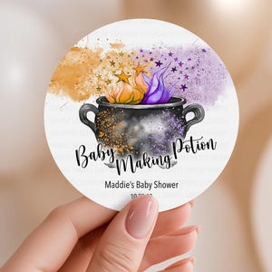May include: A round party favor sticker with a black cauldron design, the words "Baby Making Potion", and "Maddie's Baby Shower". The sticker has orange, purple, and gold accents. The sticker is available in 2" or 3" options.