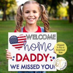 May include: A young girl holds a white sign with the words "WELCOME home DADDY WE MISSED YOU!" in red and blue. The sign features a heart with an American flag design. The background is a green grassy area.
