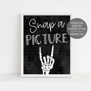 Puede incluir: Impresión enmarcada en blanco con las palabras "Snap a PICTURE" en una fuente manuscrita, y una mano de esqueleto haciendo un gesto de rock and roll. El fondo es negro texturizado. Un gráfico circular dice "Descargar ahora o enviado a usted Tabletop Sign".