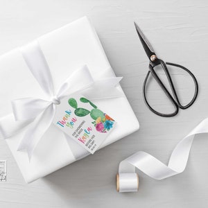 Puede incluir: Una caja de regalo blanca con una cinta de sat&eacute;n blanca y una etiqueta de agradecimiento con un dise&ntilde;o de cactus. La etiqueta dice "Gracias por celebrar a la novia Kayla" y tiene un cactus con flores.