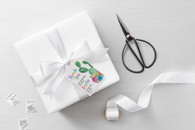 Puede incluir: Una caja de regalo blanca con una cinta de sat&eacute;n blanca y una etiqueta de agradecimiento con un dise&ntilde;o de cactus. La etiqueta dice "Gracias por celebrar a la novia Kayla" y tiene un cactus con flores.