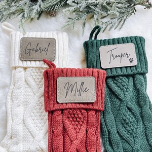 May include: Three cable-knit Christmas stockings in cream, red, and green. Each stocking has a personalized leather patch with a name: "Gabriel", "Millie", and "Trooper". The green stocking has a paw print. A festive holiday theme.