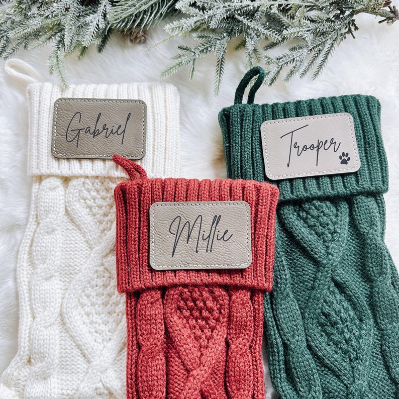 May include: Three cable-knit Christmas stockings in cream, red, and green. Each stocking has a personalized leather patch with a name: "Gabriel", "Millie", and "Trooper". The green stocking has a paw print. A festive holiday theme.