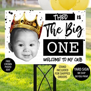 May include: Black and white yard sign with a photo of a baby wearing a gold crown. The sign says "Theo is the big one, welcome to my crib".