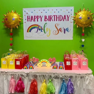 Puede incluir: Una decoraci&oacute;n de fiesta de cumplea&ntilde;os con una pancarta de arco&iacute;ris que dice "Happy Birthday Lily Sara". Hay dos globos de sol de l&aacute;mina dorada, favores de fiesta con tem&aacute;tica de arco&iacute;ris y una luz con forma de arco&iacute;ris.
