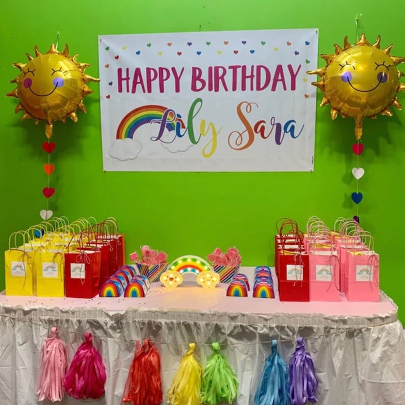Puede incluir: Una decoraci&oacute;n de fiesta de cumplea&ntilde;os con una pancarta de arco&iacute;ris que dice "Happy Birthday Lily Sara". Hay dos globos de sol de l&aacute;mina dorada, favores de fiesta con tem&aacute;tica de arco&iacute;ris y una luz con forma de arco&iacute;ris.