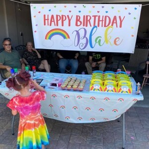Puede incluir: Una celebraci&oacute;n de cumplea&ntilde;os con una pancarta que dice "Feliz cumplea&ntilde;os Blake" con un arco&iacute;ris y nubes. Hay cupcakes en una mesa con un mantel de arco&iacute;ris y cajas de regalo para la fiesta.