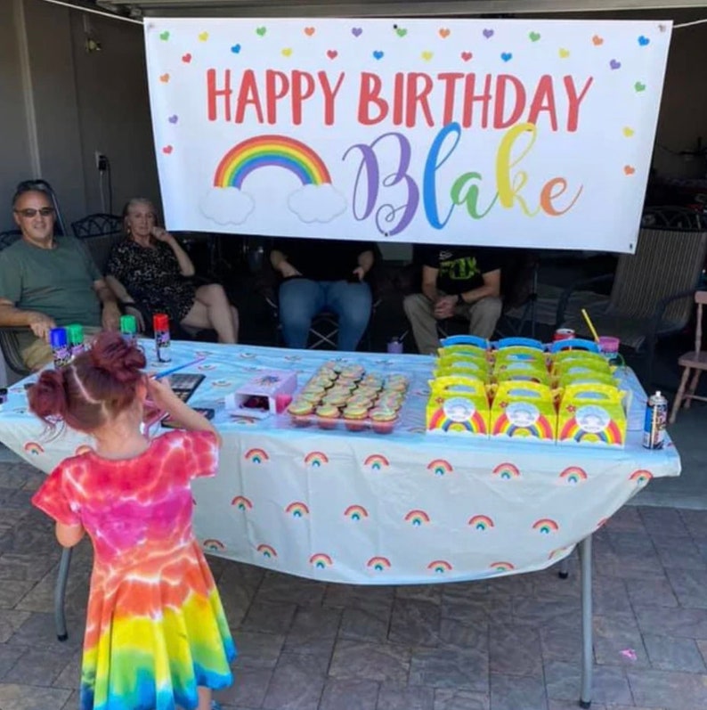 Puede incluir: Una celebraci&oacute;n de cumplea&ntilde;os con una pancarta que dice "Feliz cumplea&ntilde;os Blake" con un arco&iacute;ris y nubes. Hay cupcakes en una mesa con un mantel de arco&iacute;ris y cajas de regalo para la fiesta.