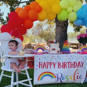 Puede incluir: Una silla alta blanca con una ni&ntilde;a peque&ntilde;a sentada en ella. La ni&ntilde;a lleva una camisa blanca y un gorro de cumplea&ntilde;os. Hay un arco de globos arco&iacute;ris detr&aacute;s de la silla alta. Una mesa con un mantel blanco y una pancarta que dice "Feliz cumplea&ntilde;os Rosalie" est&aacute; delante de la silla alta. Hay un pastel arco&iacute;ris y una torre de macarons en la mesa.