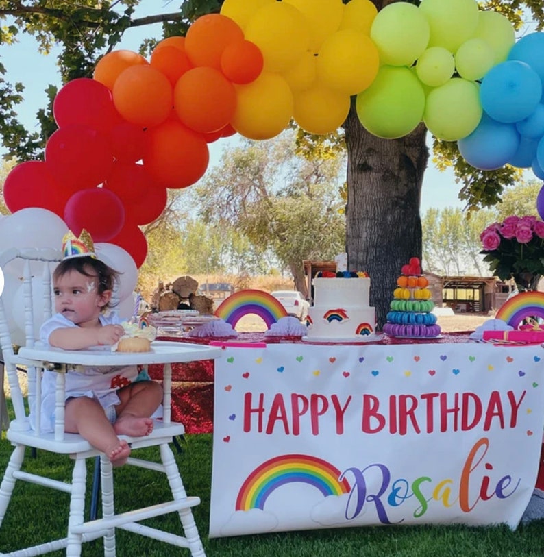 Puede incluir: Una silla alta blanca con una ni&ntilde;a peque&ntilde;a sentada en ella. La ni&ntilde;a lleva una camisa blanca y un gorro de cumplea&ntilde;os. Hay un arco de globos arco&iacute;ris detr&aacute;s de la silla alta. Una mesa con un mantel blanco y una pancarta que dice "Feliz cumplea&ntilde;os Rosalie" est&aacute; delante de la silla alta. Hay un pastel arco&iacute;ris y una torre de macarons en la mesa.