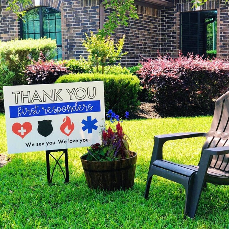 May include: A white sign with the text "THANK YOU first responders" in black lettering. The sign has a blue stripe with a red heart, a black shield, a red flame, and a blue star. The sign is on a black metal stand in a grassy yard. The text "We see you. We love you." is below the symbols.