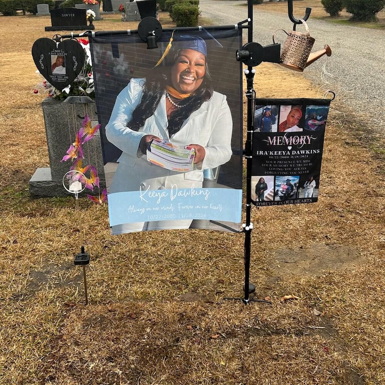 Pode incluir: Uma exibi&ccedil;&atilde;o memorial num cemit&eacute;rio. A exibi&ccedil;&atilde;o inclui uma foto grande de uma mulher com um chap&eacute;u e uma beca de formatura, com o nome "Keeya Dawkins" e uma mensagem memorial. Uma faixa menor diz "Memory" e lista detalhes adicionais.