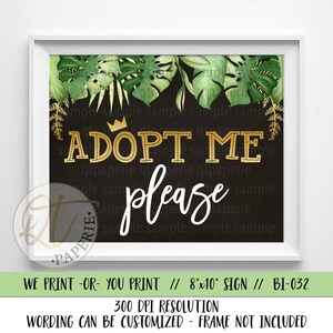 May include: A black chalkboard sign with a gold crown and the text "Adopt Me Please" in gold and white lettering. The sign is decorated with green tropical leaves. The sign is 20.32 x 25.4 cm.