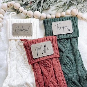 May include: Three cable-knit Christmas stockings in white, red, and green, each with a personalized leather patch. The white stocking reads "Gabriel", the red "Millie", and the green "Trooper" with a paw print. Festive holiday decor.