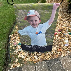 Pode incluir: Uma bandeira de jardim com uma fotografia de uma crian&ccedil;a sorrindo usando um bon&eacute; de beisebol e uma camiseta branca, com um bra&ccedil;o levantado. A bandeira tem o texto "Persy Lynn Burnette" e "We love you best friend" com datas.