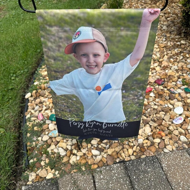 Pode incluir: Uma bandeira de jardim com uma fotografia de uma crian&ccedil;a sorrindo usando um bon&eacute; de beisebol e uma camiseta branca, com um bra&ccedil;o levantado. A bandeira tem o texto "Persy Lynn Burnette" e "We love you best friend" com datas.