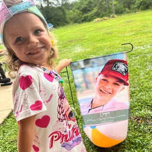 Pode incluir: Uma menina sorri enquanto segura uma bandeira de jardim com a foto de um menino usando um bon&eacute; vermelho. A bandeira tem um desenho de bola de praia na parte inferior e o texto "Cooper 2009-2024". A menina usa uma faixa de cabe&ccedil;a colorida e um vestido com estampa de cora&ccedil;&atilde;o.